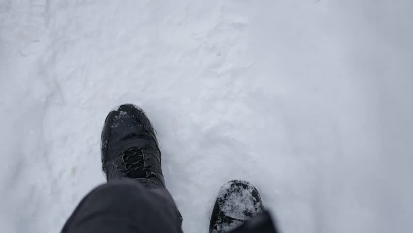 POV shot of a person walking through fresh snow in winter. Close-up of black boot stepping on white snowy ground. Cold weather, outdoor winter activity, seasonal atmosphere, slow movement in snowy landscape.