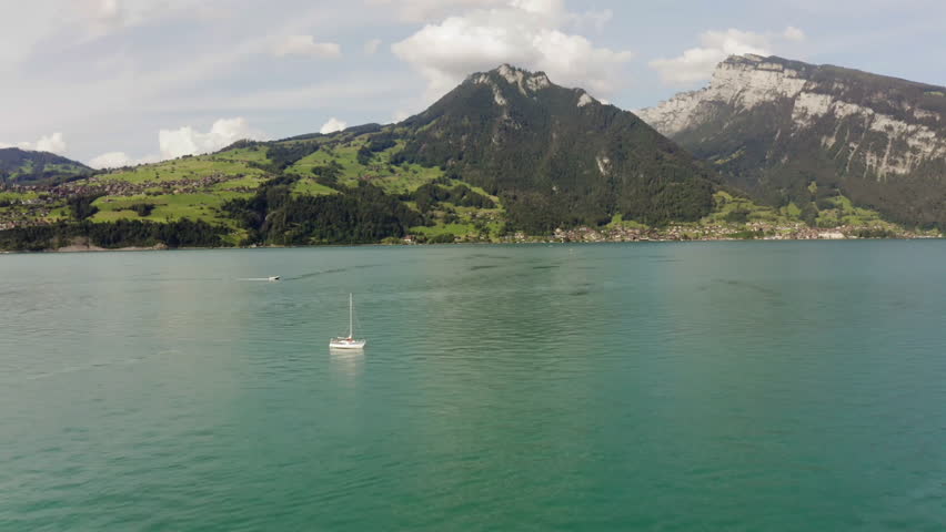A tranquil aerial shot of a lake with clear turquoise waters, bordered by lush green hills and majestic mountains under a partly cloudy sky.