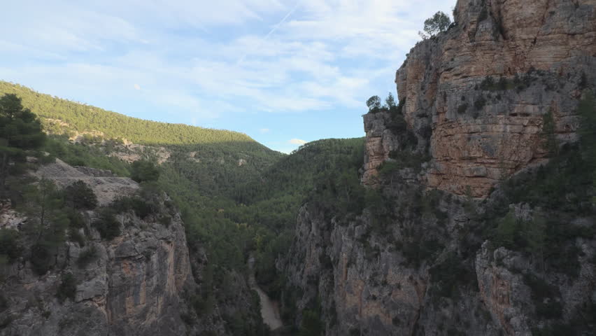 A gentle aerial glide over a limestone canyon and winding river shifting light, deep shadows, and a blue, clouded sky with a contrail evoke scale and solitude.