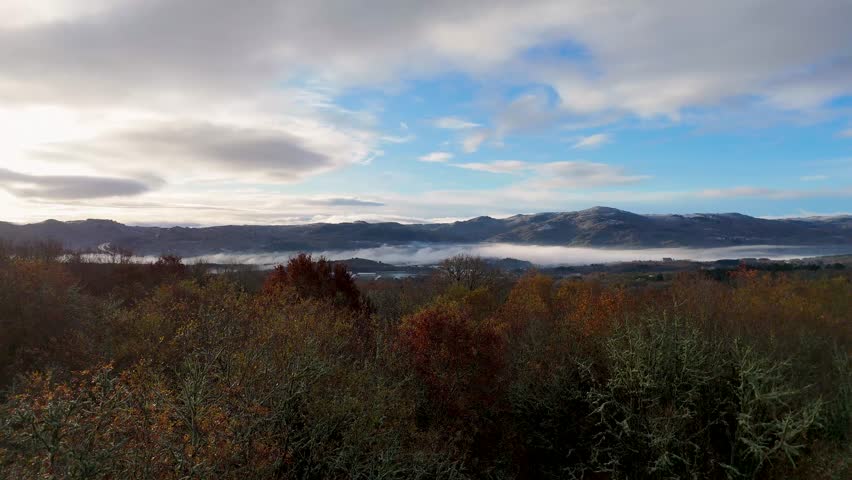 Drone flying over a foggy mountain top with a cloudy sky. The sky is blue and the mountains are covered in trees in Orense, Spain.