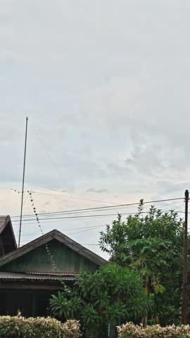 A video of a simple wooden house in rural Indonesia against a cloudy afternoon sky. Surrounded by green trees and a tranquil village atmosphere, this image showcases the natural atmosphere, the quaint village life, and the changing weather before a rainy day. The vertical composition showcases the details of the zinc roof, electricity poles, and dramatic gray clouds, perfect for content themed around rural areas, overcast weather, and simple living.