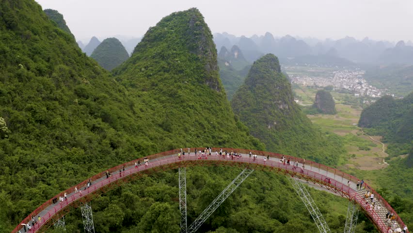 Aerial pullback shows red Skybridge curving over karst peaks in Yangshuo Gulin. Hazy valleys frame the lively span blending nature and adventure for scenic footage.