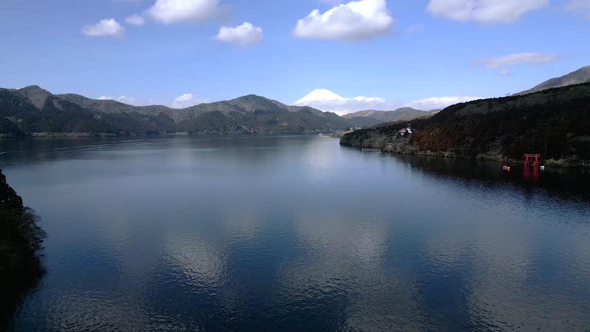 Wide panoramic view of Lake Ashi in Hakone surrounded by mountains under blue sky. Calm water with red torii gate visible on shore. Beautiful natural landscape in Kanagawa Prefecture Japan.