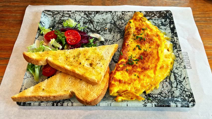 Closeup of a golden cheese omelette served with toasted bread and fresh side salad with cherry tomatoes. Casual lunch plate in a cozy restaurant setting.