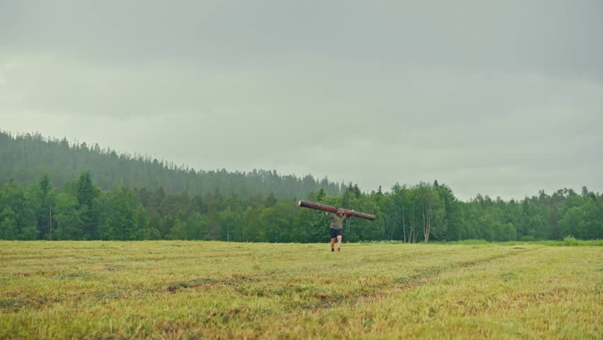A Man Carrying A Log Over His Shoulders Walking Across The Fields. Wide Shot