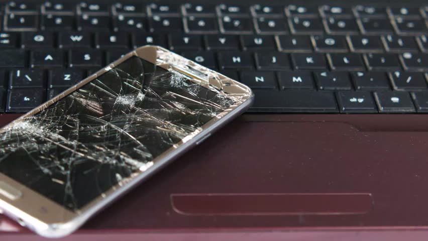 A close up view of a broken smartphone with a shattered screen resting on a dark red laptop keyboard