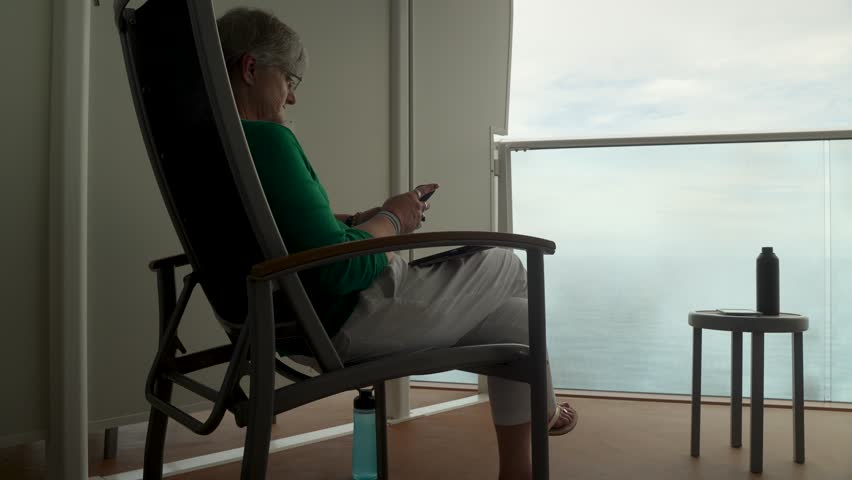 A senior woman relaxes on a cruise ship balcony using her smartphone. The serene sea view sets a tranquil mood. Soft lighting enhances a cozy atmosphere against the water backdrop.