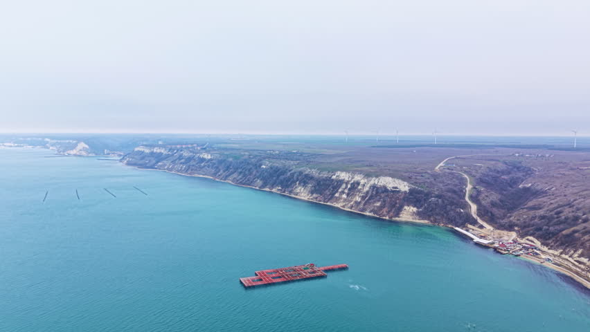 A mussel farm sits in the sea, with floating structures used for cultivation. Land and water meet at the coast, while distant works and wind turbines appear in the background.