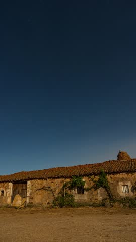 Stunning timelapse showing stars creating circular trails in the deep blue night sky over a rustic, abandoned stone farmhouse in the countryside