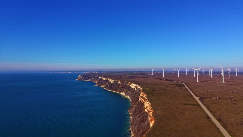 Wind turbines stand tall along the coastline with cliffs rising above the water. The clear sky enhances the view of the land meeting the sea in the background.