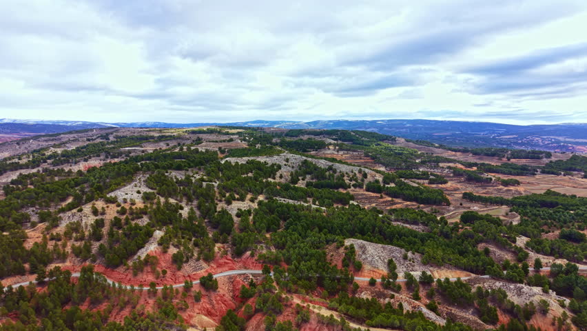 A wide view of hills covered with trees and patches of red earth. Winding roads can be seen connecting different areas in this rural landscape under a cloudy sky.