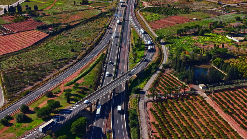A view shows a busy highway with cars and trucks traveling. Fields and green areas surround the road on both sides. The scene is bright with clear skies.