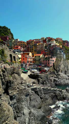 Aerial drone pan across Riomaggiore in Cinque Terre, Italy, showing colourful cliffside houses above the rocky harbour and Mediterranean Sea on a sunny summer afternoon.
