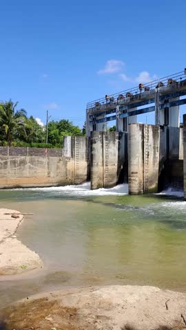 Vertical video for mobile phone screens. A powerful dam structure on a bright day. The image features water cascading through the dam and flowing over a sandy shore