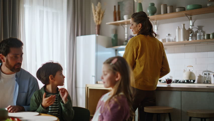 Caring mom serving table preparing breakfast dining room closeup. Smiling mother putting plates kissing kids. Friendly people spending weekend cozy light apartment. Parents children home morning