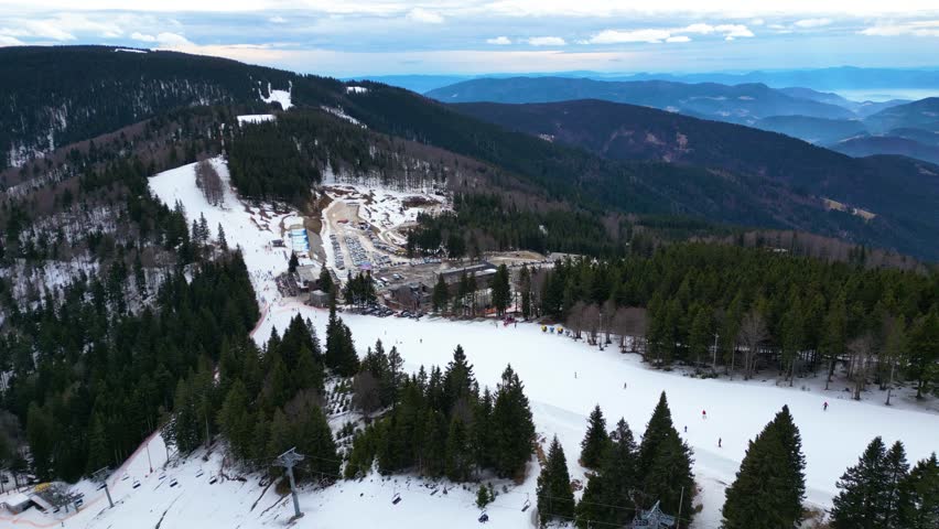 Aerial view of ski slope and chairlift at Kope ski center, Slovenia, with skiers on groomed piste, surrounded by forest and scenic winter mountains.