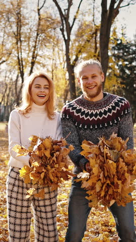 Autumn fun. Young happy man and woman throwing bunches of fallen leaves in park and laughing under rain, fooling around together, slow motion
