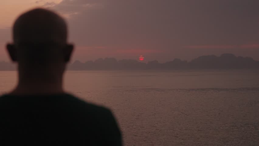 Silhouetted tourists watch the sun set over Ha Long Bay, Vietnam, as warm light reflects on calm water and limestone karsts. A serene evening travel scene capturing the bay’s iconic atmosphere.