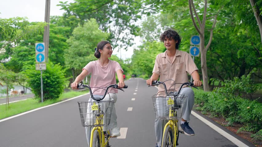Asian young couple dating and biking together on a road in the park. Attractive man and woman feel happy and enjoy riding bicycles side by side, spend free leisure time and outdoors activity in garden
