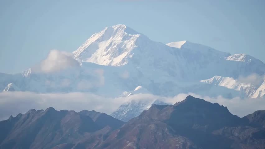 Snowcapped Denali rises above a sea of clouds, its towering peak gleaming in sunlight over vast Alaskan wilderness and dramatic alpine skies.