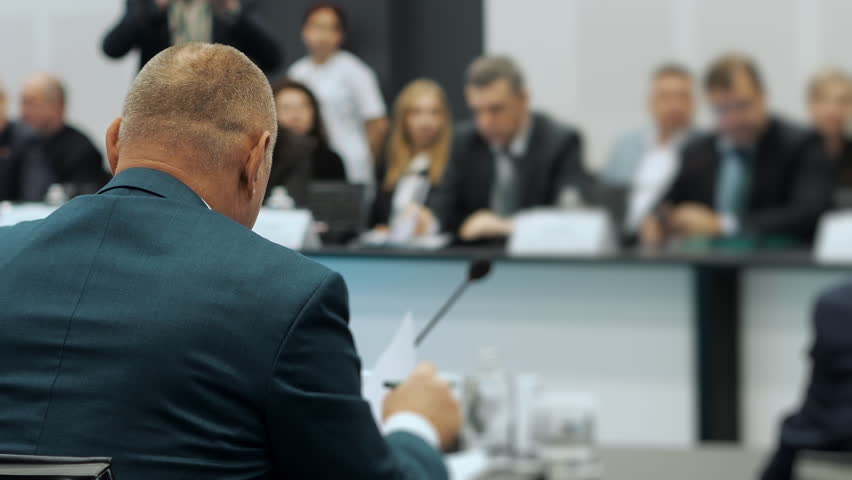 Mature businessman in suit speaking at formal board meeting in modern conference room, corporate leadership discussion and decision making indoors.