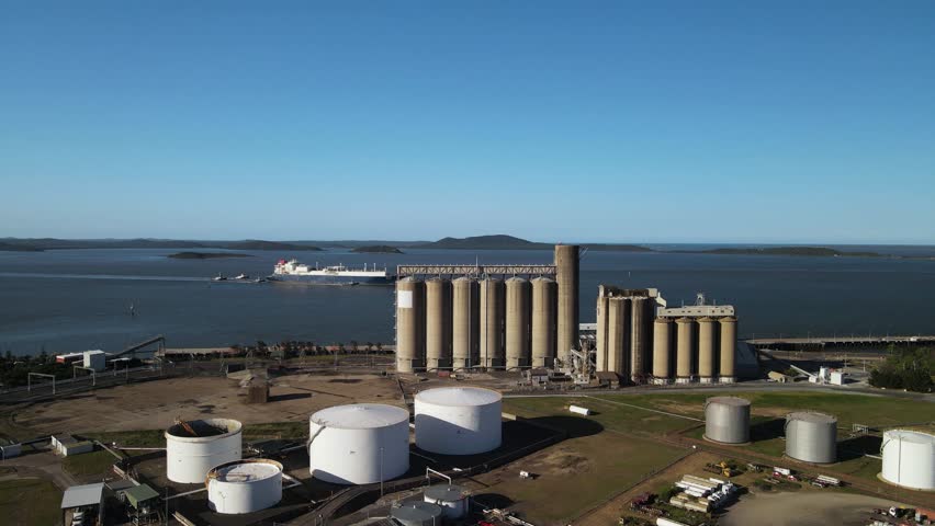 A large cargo ship navigates the waters of a industrial port near a towering storage harbor facility. Moving drone view