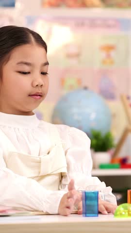 Thoughtful elementary girl studies colorful geometry blocks at classroom desk, soft lighting, static medium shot