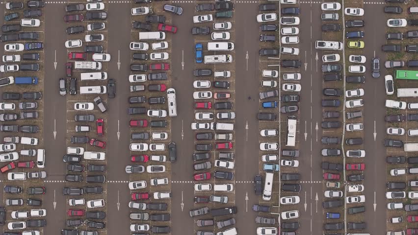 Overhead aerial perspective reveals hundreds of densely parked passenger cars trucks and vans occupying every available space within the sprawling outdoor parking facility delineated by white painted lines.
