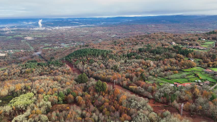 Drone flying over a beautiful view of a forest with a few houses in the distance. The trees are full of leaves and the sky is cloudy in Orense, Spain.