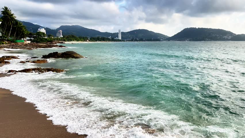 A peaceful beach in Phuket, Thailand. Gentle waves wash up on the shore with green hills and modern buildings in the background, creating a tropical vacation atmosphere.