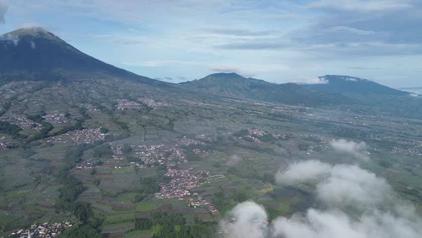 View of Mount Sindoro with fluffy clouds and rural rice field atmosphere recorded from the foot of the mountain using a drone