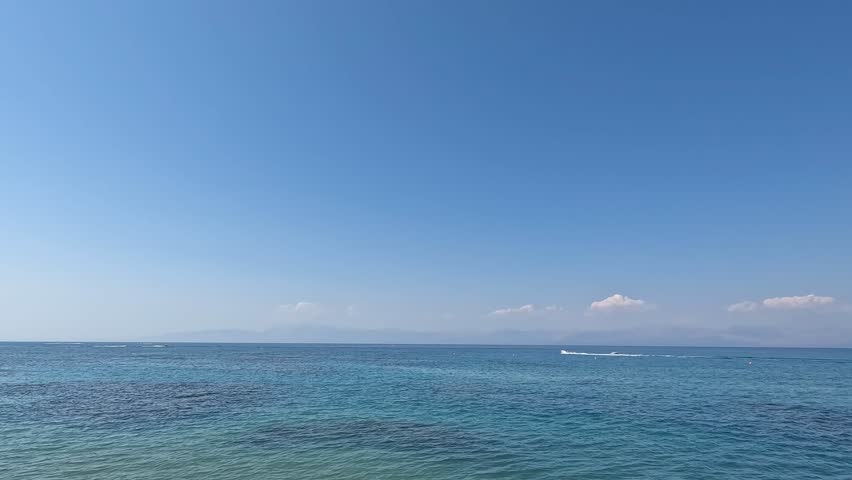 A wide panoramic view of the calm, turquoise Ionian Sea under a clear blue sky, with the hazy outlines of coastal mountains in the far distance.