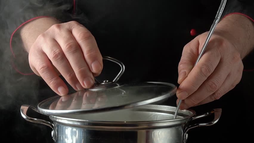 Chef in black uniform lifts lid of pan, stirring pasta with ladle, steam rising in bright restaurant kitchen environment.