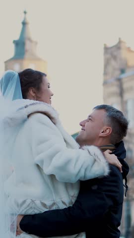 winter wedding. newlywed couple in wedding dresses. groom holds bride in his arms, spinning. they are happy, smiling at each other. background of ancient architecture, snow-covered park,