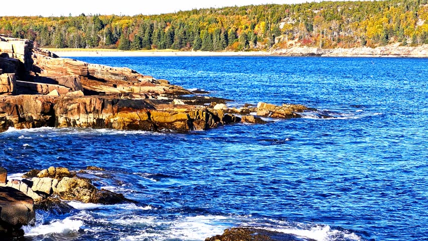 Scenic view of the Atlantic ocean crashing on the rocky coast of Maine. Beautiful autumn foliage covering the landscape on a sunny day with Sandy Beach in distance.