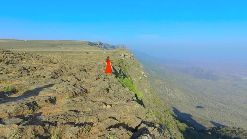 Aerial view of a young woman in a red dress standing on the rocky cliff edge of the plateau at Jabal Samhan, Salalah, Oman.