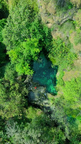 Aerial view of a young woman in a red dress sitting on a rocky ledge overlooking a turquoise sinkhole surrounded by trees in Salalah, Oman.