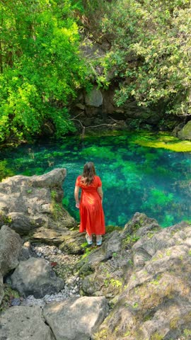 Aerial view of a woman in a red dress standing on rocky cliffs overlooking a clear green natural spring in Salalah, Oman.