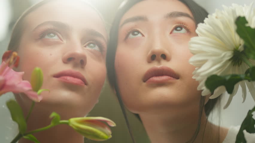 Two diverse women looking up at bright sunlight with blooming flowers indoors Asian Caucasian girls female dreamy hopeful faces smooth skin natural beauty harmony spring fresh seasonal atmosphere