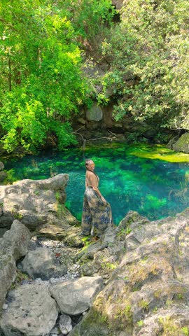 Aerial view of a young woman standing beside a clear turquoise water pool surrounded by lush green trees and rock formations in Salalah, Oman.