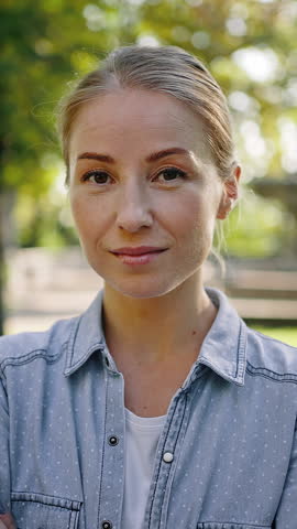 Young woman in denim jacket looks in camera smiling on blurred background. Blonde lady stands in sunny city park against green trees closeup