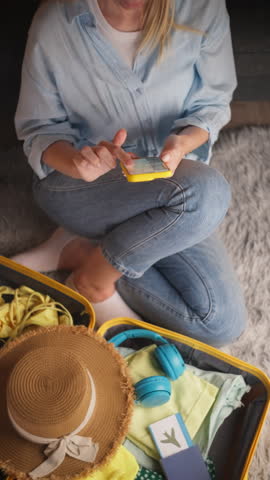 Young traveler woman sitting on floor using mobile phone to search for holiday destinations. Booking tickets and hotel online with an open suitcase full of summer essentials ready for trip