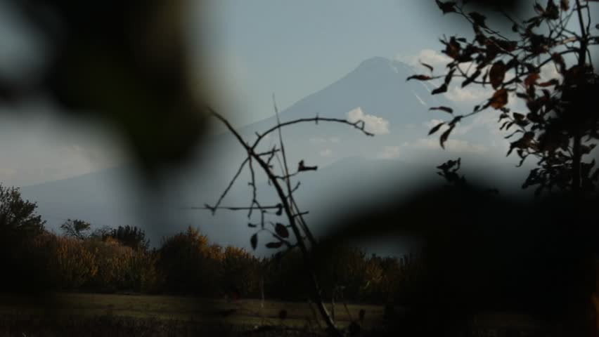  A serene view of a majestic, snow-capped mountain peak rising above misty layers and soft clouds, set against a clear sky with autumn trees in the foreground.