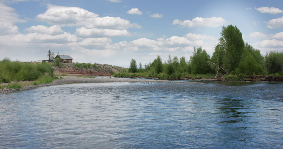 Summer footage of a peaceful float down the Green River just outside of Pinedale, Wyoming. The video shows calm river waters, cottonwood trees lining the shoreline, and a remote cabin visible in the distance, capturing the quiet rural beauty of the American West. Ideal for travel, tourism, outdoor recreation, fly fishing, nature, and lifestyle stock footage.
