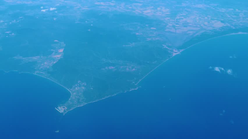 coastal landscape curved peninsula deep blue waters faint urban clusters trace shoreline under hazy high altitude broad aerial view sea atmosphere air travel