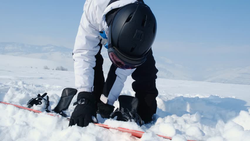 Snowboarder lifts snowboard from snow and shakes off stuck snow, cleaning board surface. Mountain winter sport, passion for snowboarding on frosty winter day.