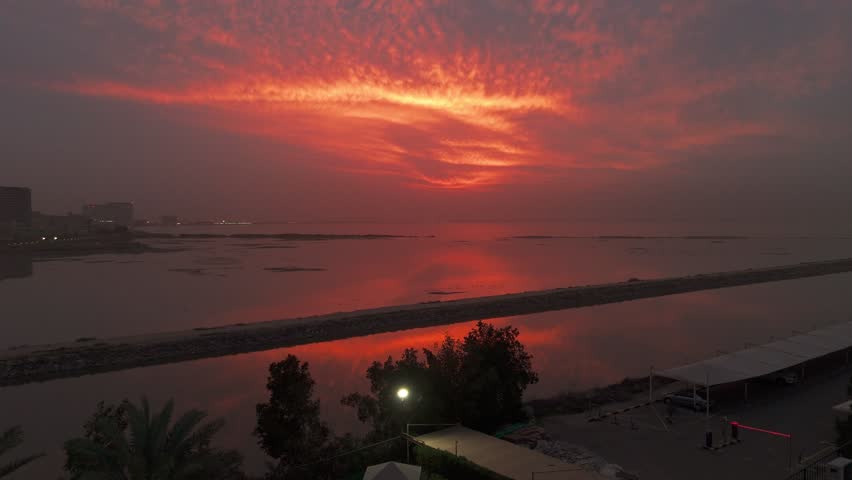 Wide view of fiery red twilight clouds reflecting on calm lagoon water, with dark sandbar shoreline and distant buildings creating a moody coastal panorama.