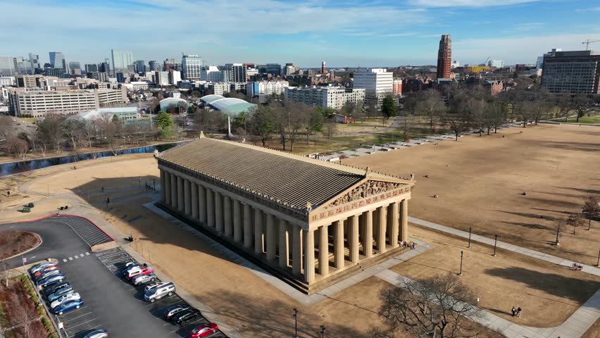 Aerial view of Parthenon in Nashville, Tennessee with downtown skyline in background. Historic landmark in Centennial Park under clear sunny sky with urban cityscape. Wide shot. Approaching.