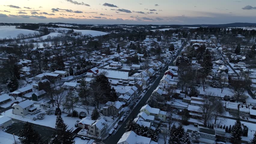 Drone flight over snowy winter city in America with Main Street at dusk. Aerial wide shot. Scenic rural landscape in distance. Residential area or neighborhood in USA. White winter wonderland.