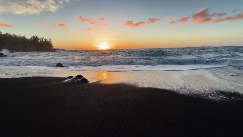 Strong ocean waves rolling onto black sand beach at sunrise in Hawaii